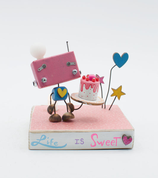 Diorama scultura luminosa stile robot fatto a mano rosa con testa bianca e cuore giallo che tiene torta su vassoio in legno