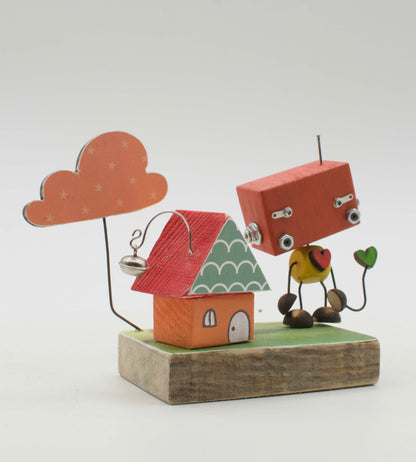 Diorama scultura luminosa in legno fatta a mano con robot rosso e giallo accanto a casetta miniatura su base rustica