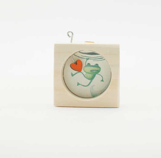 Piccolo quadro artigianale in legno con inserto di vetro circolare raffigurante una rana verde che tiene un cuore arancione